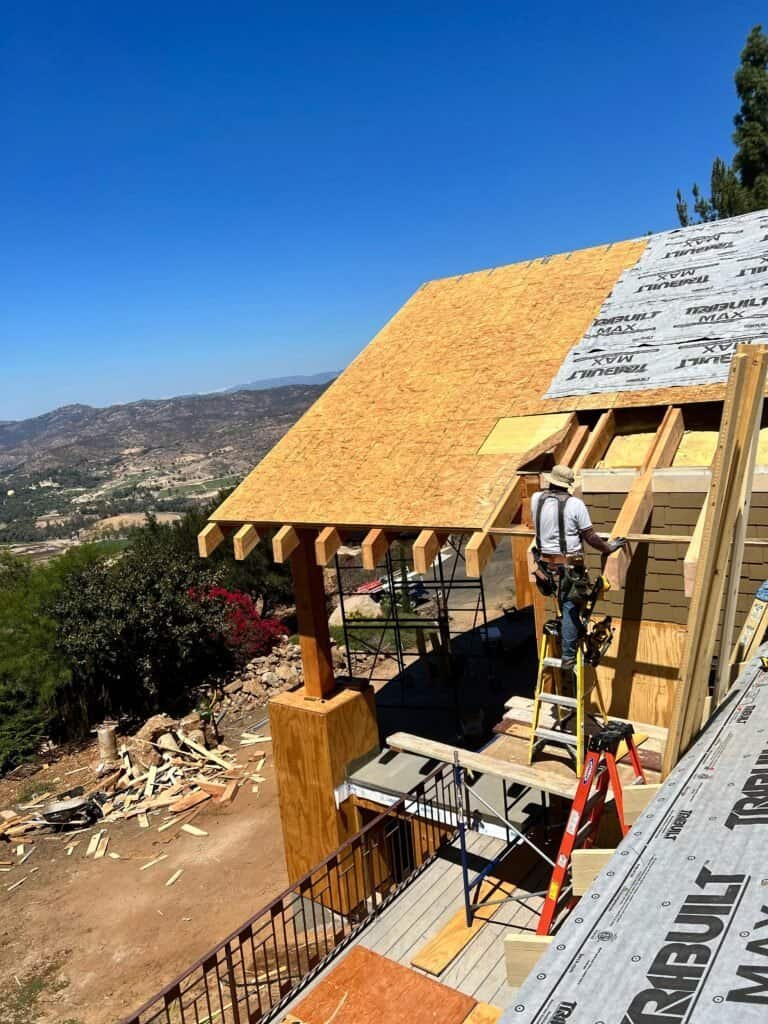Wooden house roof construction under clear blue sky with mountain view, ongoing building process, framing and roofing work by contractors.